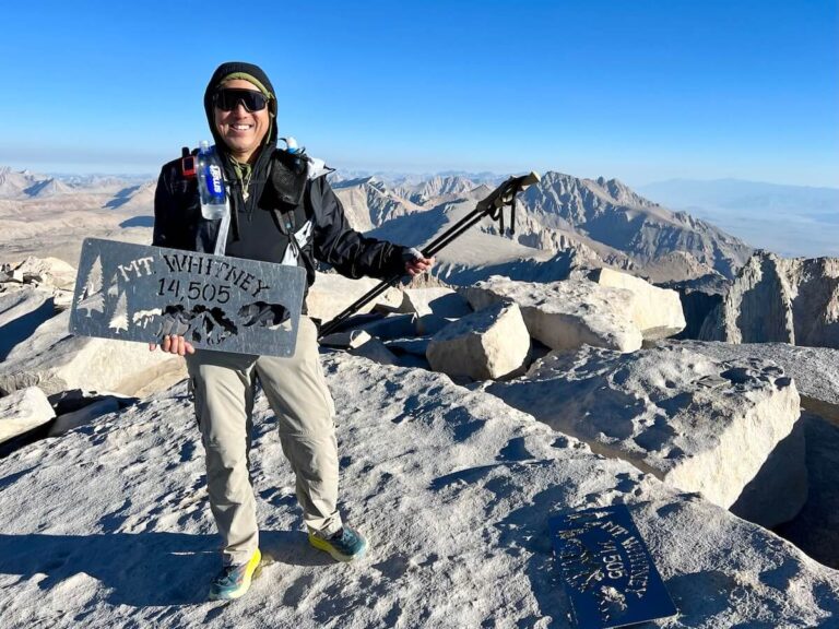 Jeff on the summit of Mount Whitney, holding the metal summit sign at 14,505 feet. His backpacking essentials are visible, including the Garmin InReach Mini 2, Zpacks Arc Haul Ultra, HOKA Speedgoat w/ Vibram shoes, Gossamer Gear LT5 Three Piece Carbon Trekking Poles, Smith Wildcat Glasses, Outdoor Vitals Ventus Hoodie, Patagonia Sun Hoodie, Columbia Silver Ridge Utility Convertible Pants, Katadyn BeFree water filter with HydraPak bottle in a Zpacks Water Bottle Sleeve, and Outdoor Research Sun Gloves.