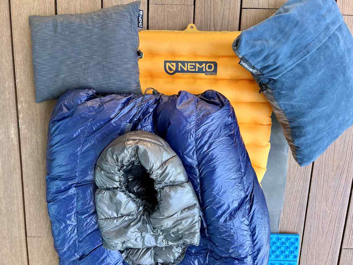 Close-up of a Nemo Tensor insulated pad with an Enlightened Equipment Revelation 20-degree quilt, Gossamer Gear 1/8-inch Thinlight pad, Zpacks Z-Seat, Nemo Fillo Elite pillow, and Therm-a-Rest Compressible Pillow laid out for a backpacking setup