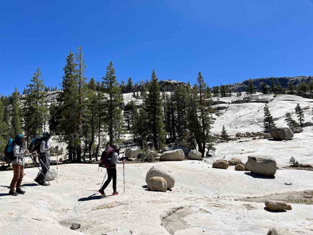 Group of hikers traversing granite slabs in Yosemite National Park, approaching Polydome, surrounded by pine trees and clear blue skies.