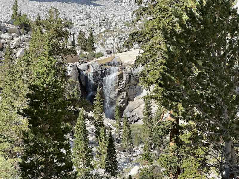 A hidden waterfall tumbles over granite cliffs behind Outpost Camp on the Mount Whitney Trail, framed by evergreen trees and rugged high Sierra landscape.