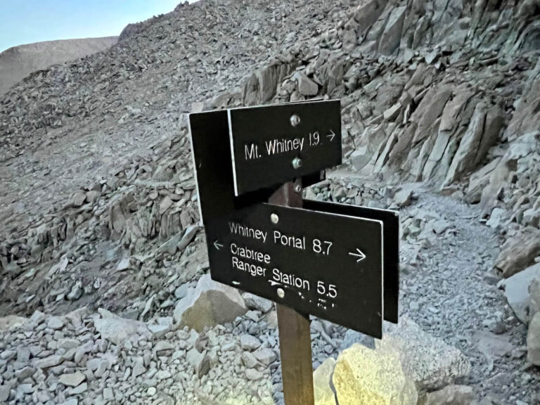 Trail sign showing directions to Mt. Whitney, Whitney Portal, and Crabtree Ranger Station against rocky mountain terrain.