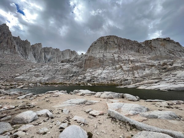 At a Mount Whitney elevation of 12,000 feet, the peaceful tarn at Trail Camp on the Mount Whitney Trail, surrounded by rugged granite cliffs under a cloudy sky.