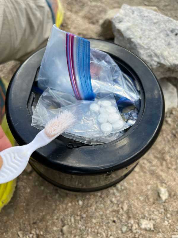 Bare Boxer Bear Canister with essentials inside, including a toothbrush and tablets, on the ground during a camping trip.