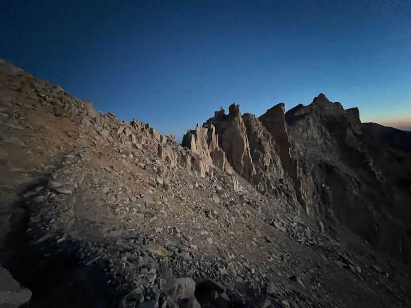 Rocky terrain near Trail Crest on Mount Whitney, with jagged peaks silhouetted against a deep blue pre-dawn sky.