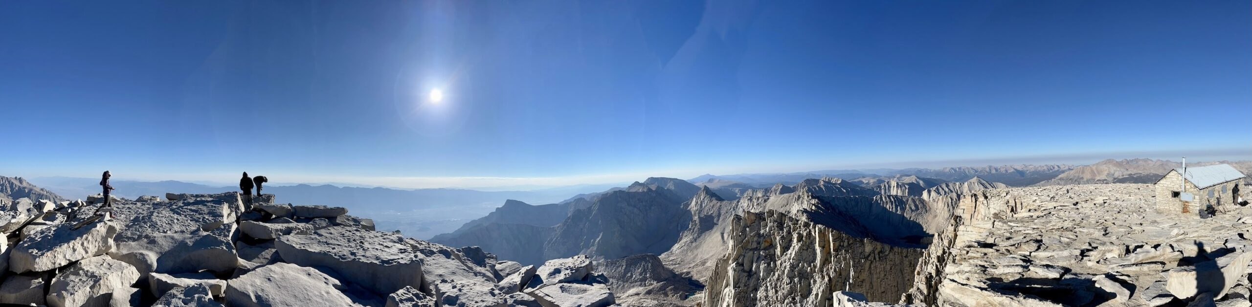 Panoramic view of Mount Whitney’s summit showing the summit blocks, the stone summit hut, the Needles, and Lone Pine Peak in the distance under a clear blue sky. Only a few climbers are visible on the rocky terrain, emphasizing the serenity of an early alpine start.