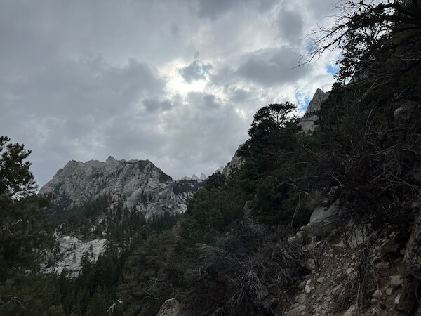 Dark clouds forming over the trail between Lone Pine Lake and the Whitney Portal, with rugged cliffs and pine trees along the rocky slope.