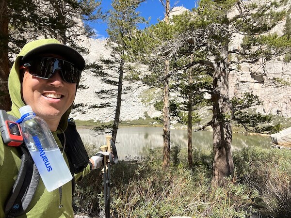 Jeff standing near the edge of Mirror Lake, with the reflections of granite cliffs and surrounding greenery visible in the water’s surface. Jeff is wearing a green Pagaonia sun hoodie, Smith Wildcat sunglasses, Garmin InReach Mini 2, Zpacks Arc Haul Ultra 60L, OR sun gloves, and 2 Gossamer Gear LT5 carbon trekking poles.