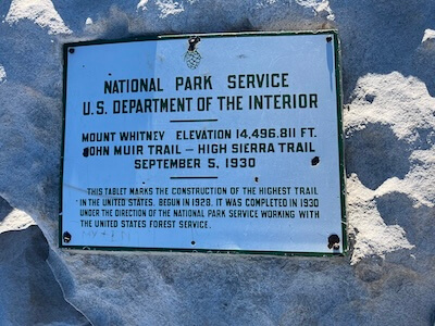 Iconic Mount Whitney summit plaque installed by the National Park Service, dated September 5, 1930, indicating an elevation of 14,496.811 feet. The plaque commemorates the construction of the highest trail in the United States at the time. The plaque is mounted on a granite rock, surrounded by rugged stone textures.