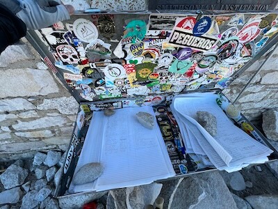Sticker-covered metal summit registry box at the Mount Whitney summit, filled with loose summit registry pages secured by rocks to prevent the pages from blowing away. Each summiteer records their name, creating a lasting entry in the summit’s history.