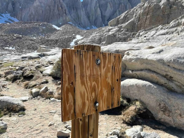 Wooden trail sign at the entrance to Trail Camp on the Mount Whitney Main Trail, weathered and nearly unreadable. The only photo I have of this sign is from my previous attempt 2.5 months earlier, taken on May 31, 2022. Snow patches in the background reflect the early-season conditions.