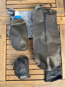 Katadyn BeFree water filter attached to a HydraPak Flux 1L bottle, alongside a HydraPak Seeker 4L water storage bag. A collapsed Seeker 4L bag is also shown to highlight its compact form factor, with a Zpacks Medium-Plus Dry Bag in the background. This setup represents a lightweight, collapsible water filtration and storage system for backpacking.
